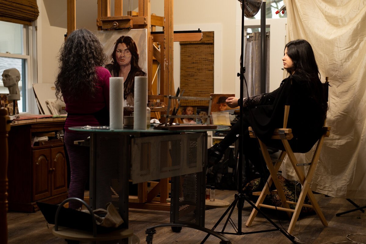 Hilarie Couture, left, paints Malak El-Taleb of Libyan heritage, right, in her home studio for her exhibit Unity with Variety.